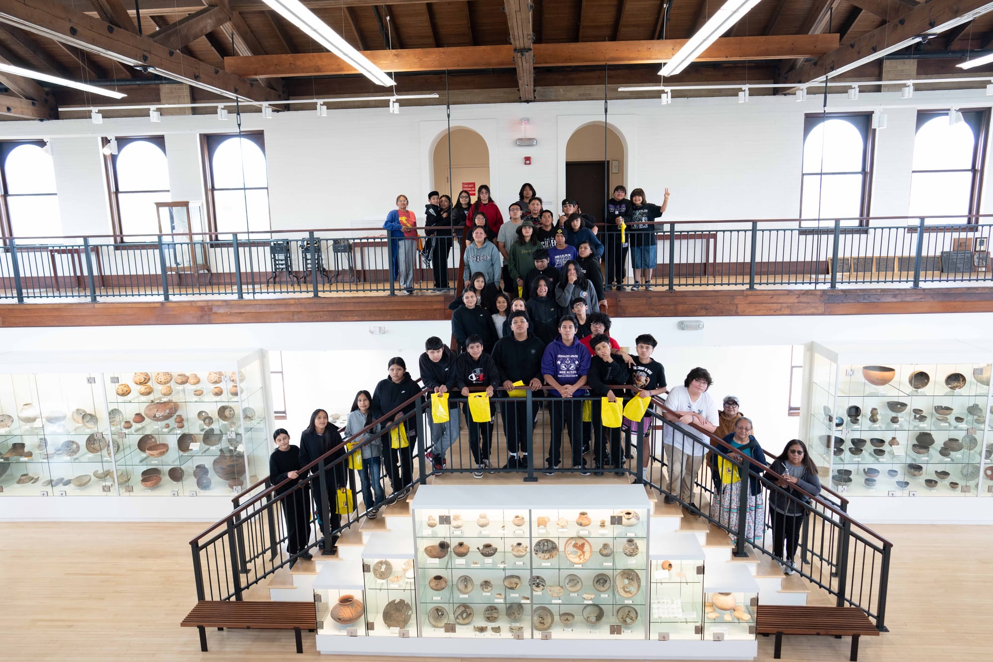 A large group of Mescalero Apache students and staff pose on a staircase and balcony inside the WNMU Museum, surrounded by display cases containing pottery and other artifacts.