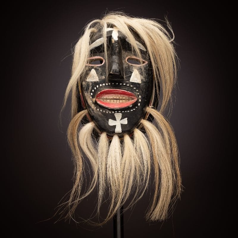 A black Mexican festival mask with blond hair, red lips, white markings and long hair is displayed against a dark background.
