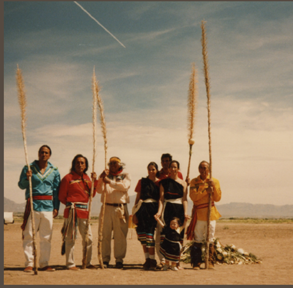 Members of the Piro-Manso-Tiwa Indian Tribe stand outdoors holding long ceremonial staffs in a desert landscape.