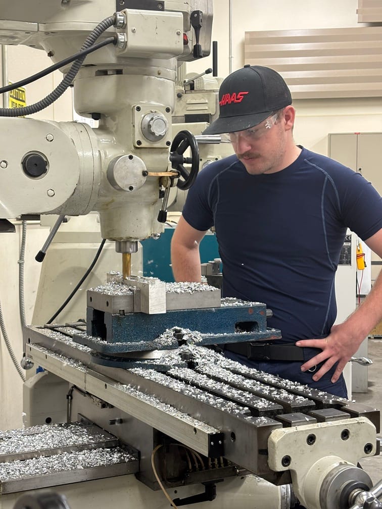 A DACC manufacturing student wearing safety glasses operates a milling machine surrounded by metal shavings.