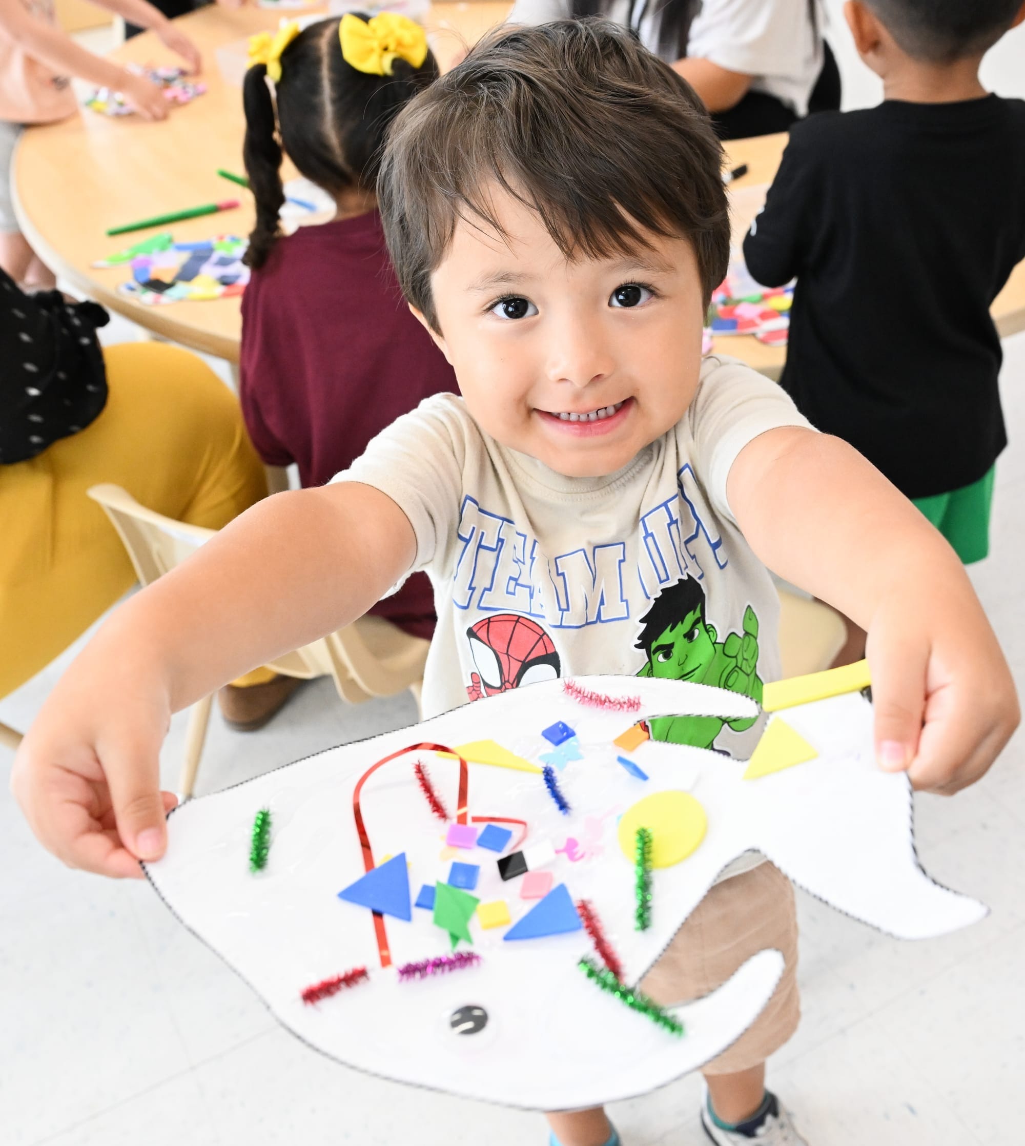 NMSU Head Start student Perseus Walker holds up a colorful art project in a classroom.