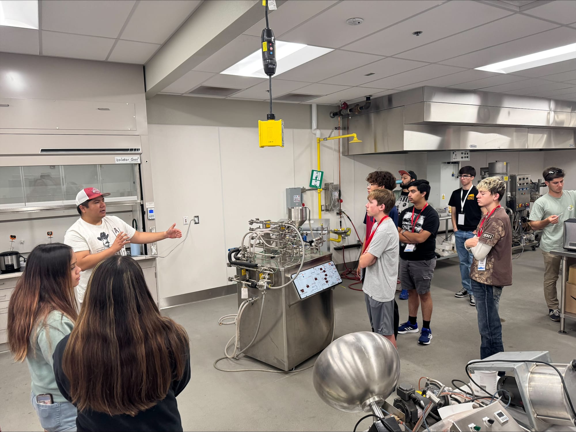 High school students attending NM PREP Academy are gathered around a machine in a chemical engineering lab while listening to an instructor.
