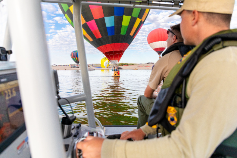 A New Mexico State Parks marine enforcement officer patrols Elephant Butte Lake by boat as hot air balloons float over the water.