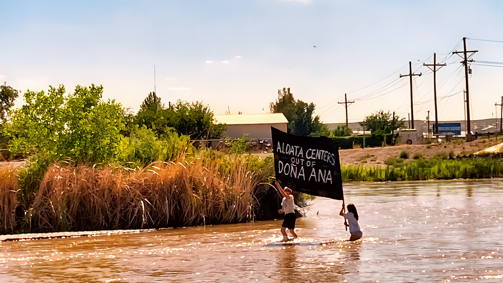 Two protesters stand in the Rio Grande holding a black banner opposing data centers in Doña Ana County.