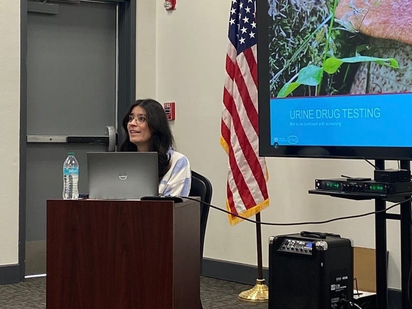 Dr. Francheska Gurule sits at a lectern during a presentation beside a screen showing information about urine drug testing.