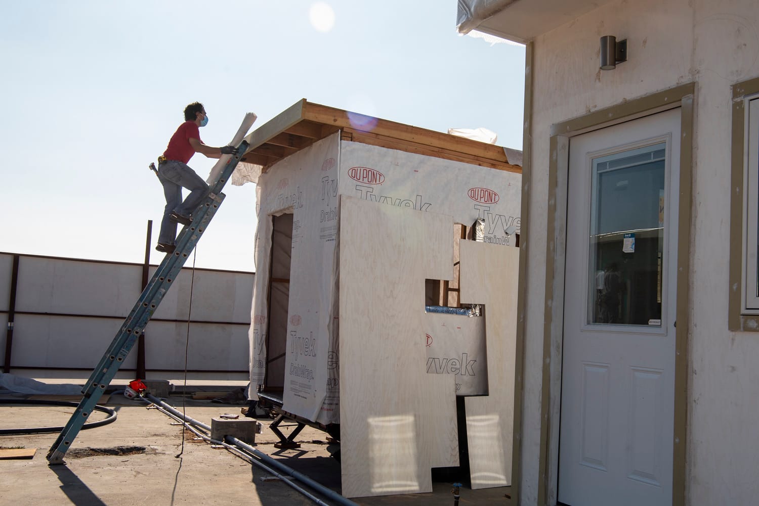 A worker stands on a ladder beside a small home under construction, with another completed small structure visible nearby.
