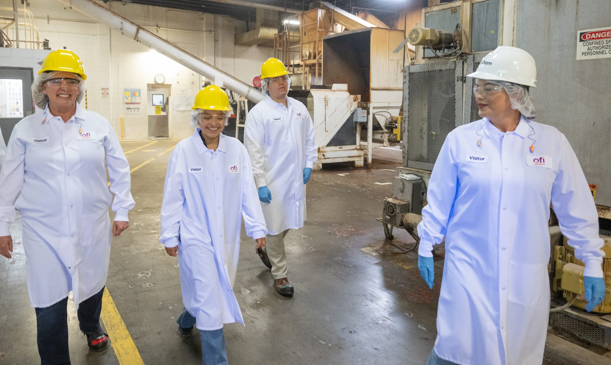 Four people wearing protective coats, hair coverings, safety glasses and hard hats walk through an Olam Food Ingredients production facility.