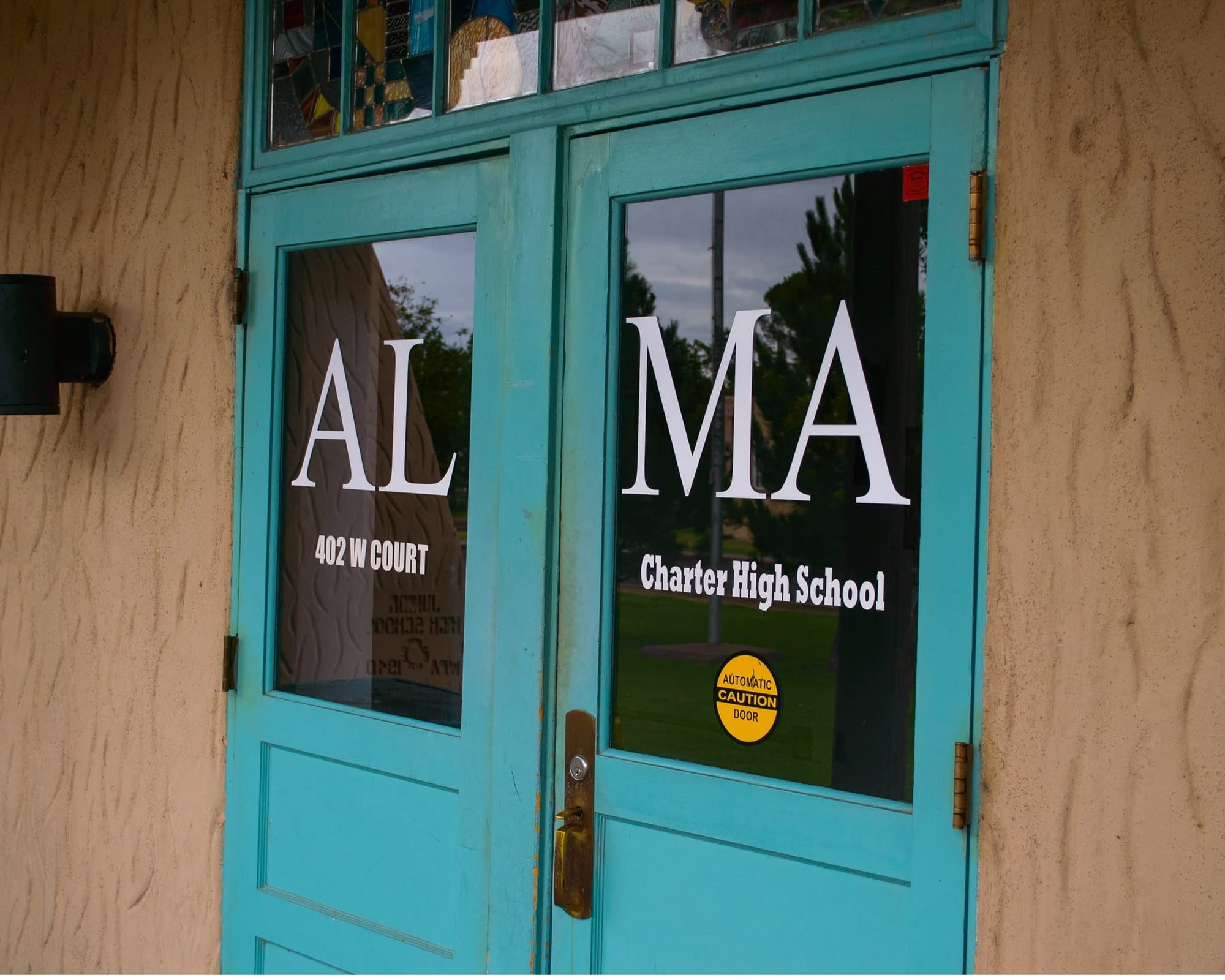 Turquoise double doors at Alma d’Arte Charter High School in Las Cruces display the school's name and 402 W. Court Ave. address in white lettering.