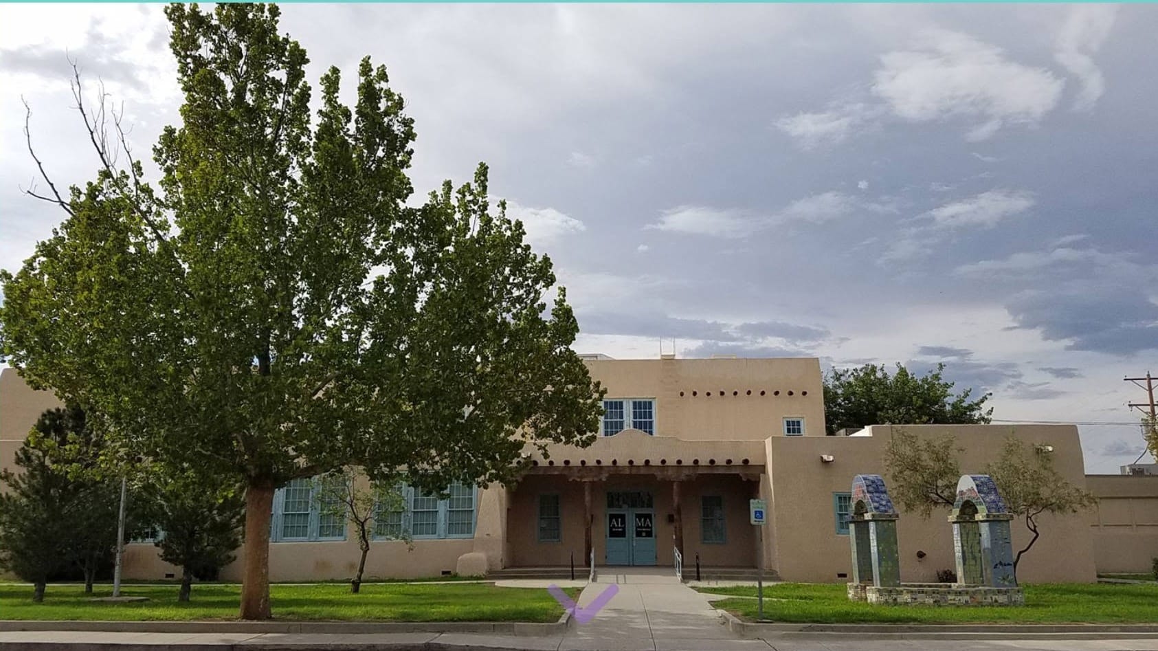 Exterior of Alma d'Arte Charter High School in Las Cruces, showing its adobe-style building, central entrance, large trees and decorative tiled arches.