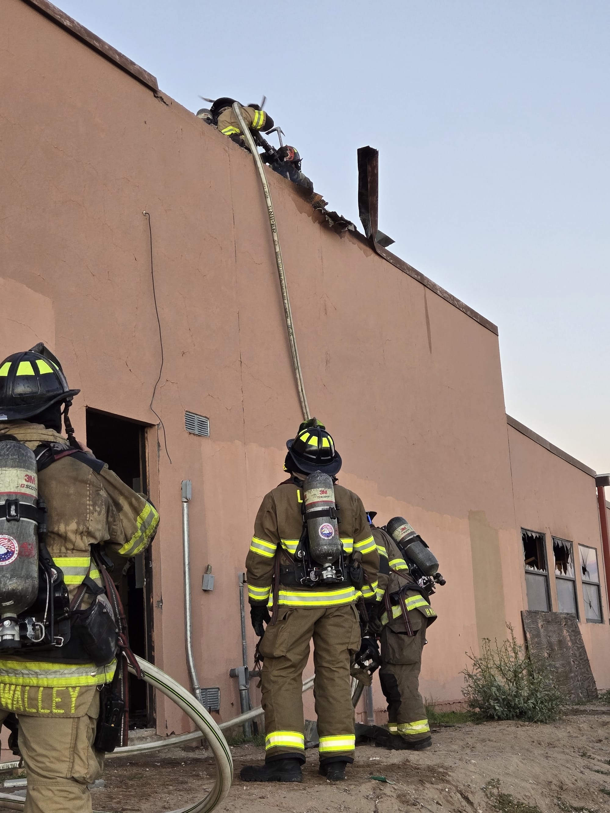 Firefighters in protective gear work from a ladder and the ground beside a fire-damaged commercial building with broken windows.