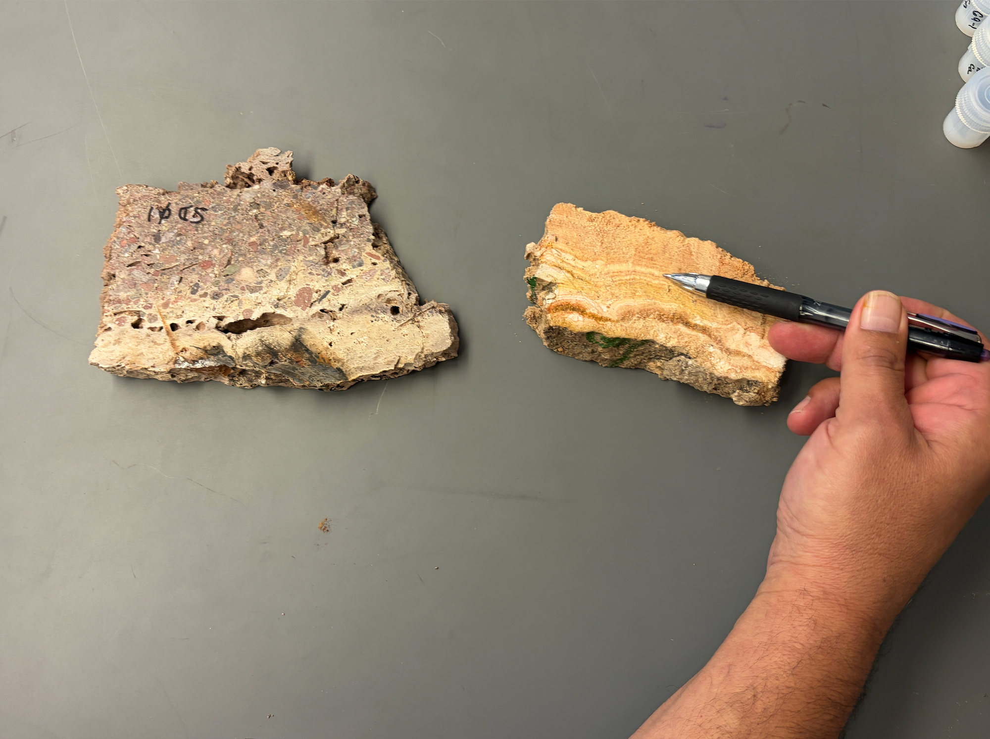Two travertine rock samples on a laboratory table at NMSU, with a hand holding a pen to point out mineral layers in one sample.