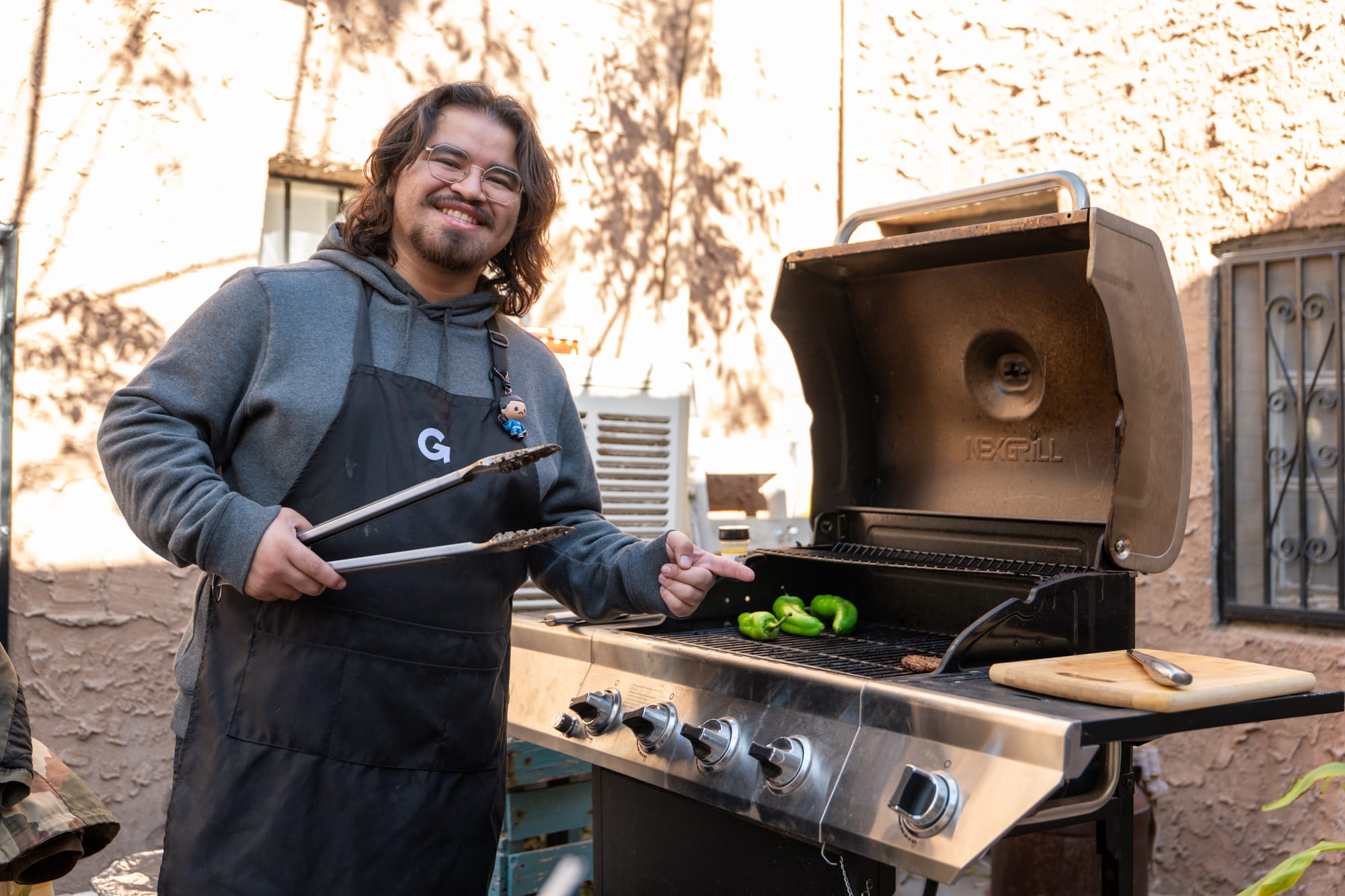 Angel Muñoz, property master and editor of “La Carne Asada,” stands beside an outdoor grill holding tongs as green chiles roast.