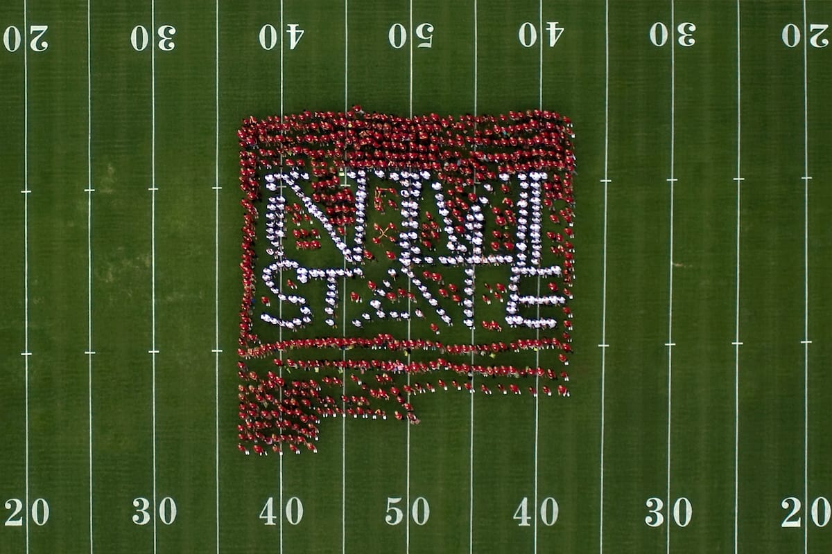 Aerial view of a large group dressed in crimson and white forming the NM State logo at midfield inside Aggie Memorial Stadium.