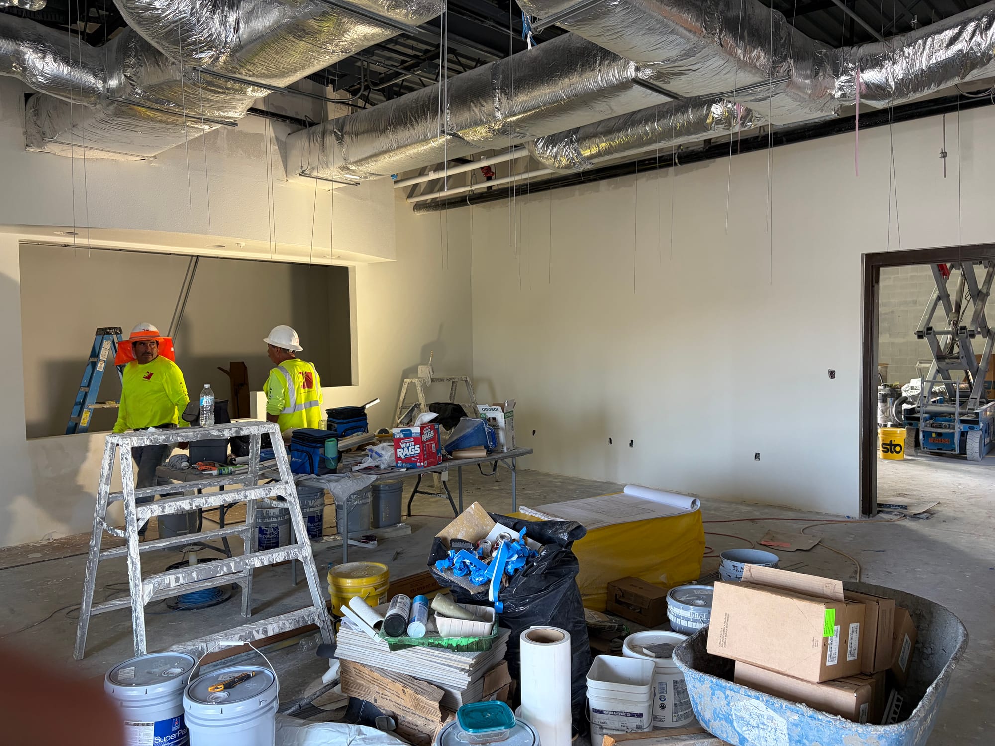 Two construction workers in high-visibility shirts and hard hats stand inside Sunland Park's new food pantry amid ladders, building supplies and exposed ceiling ductwork.