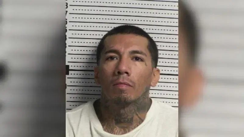 Booking photo of Ruben Jose Trujillo wearing a white shirt, with short dark hair and extensive neck tattoos, against a height-measurement backdrop.