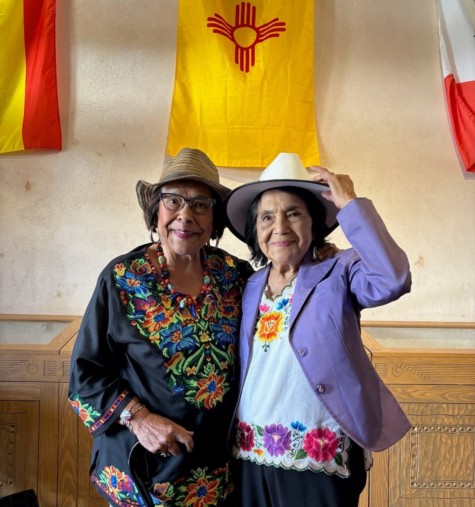 New Mexico Rep. Patricia Roybal Caballero stands beside Dolores Huerta indoors beneath hanging flags in an undated photo.