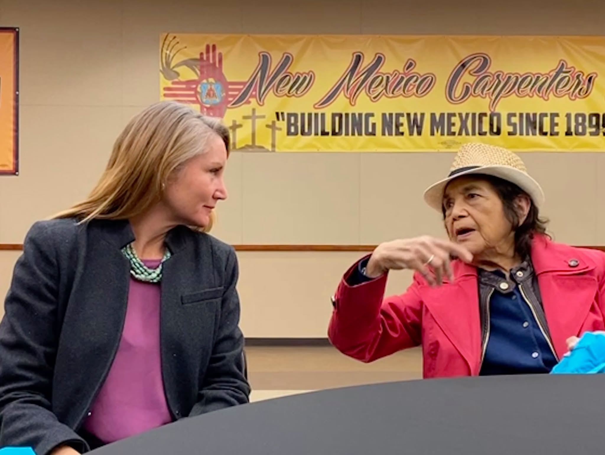 U.S. Rep. Melanie Stansbury listens as Dolores Huerta, wearing a red jacket and straw hat, speaks at a table beneath a New Mexico carpenters banner.