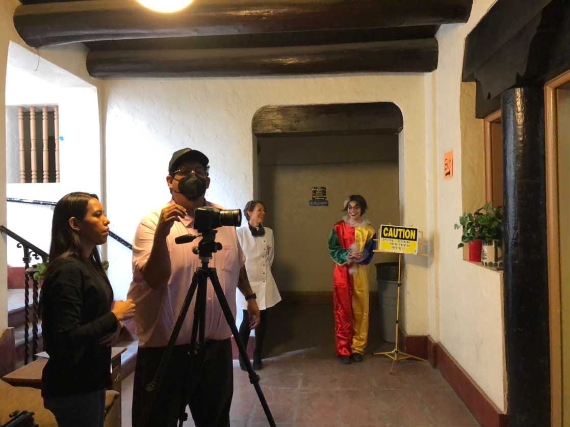 A film crew operates a camera inside the former Doña Ana County courthouse as performers in a white coat and colorful clown costume stand nearby.