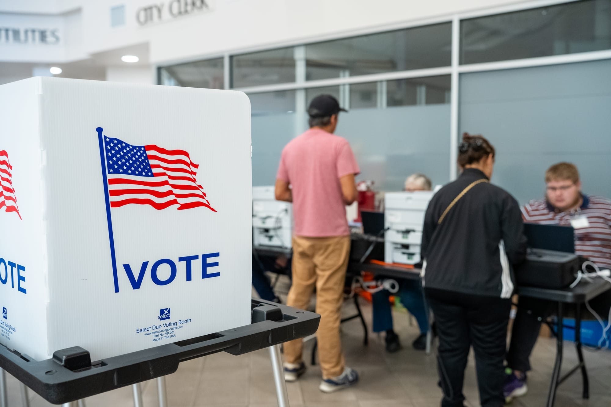 A voting booth displaying an American flag and the word “VOTE” stands in the foreground as voters check in with election workers at a Doña Ana County polling location.