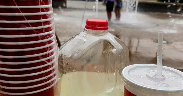 Raising Cane’s cups and lemonade jug at a previous Lunchbox Lemonade event on Plaza de Las Cruces, with children playing in the splash pad behind