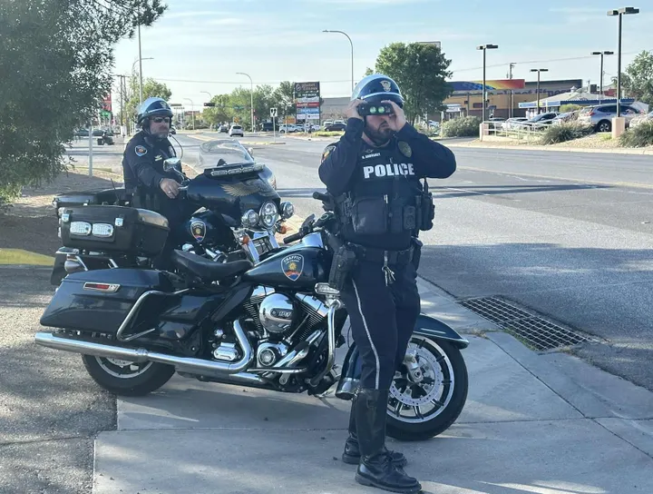 Two Las Cruces police officers on motorcycles conduct a traffic enforcement operation, with one officer using binoculars to monitor vehicles along El Paseo Road