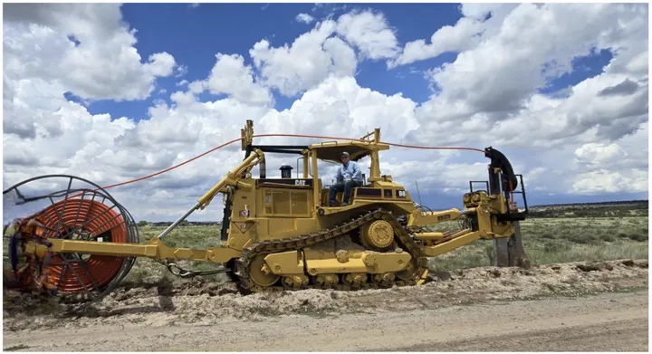 A large yellow Caterpillar D8 bulldozer pulls and buries a spool of orange fiber-optic cable along a dirt road under a partly cloudy sky in rural New Mexico.