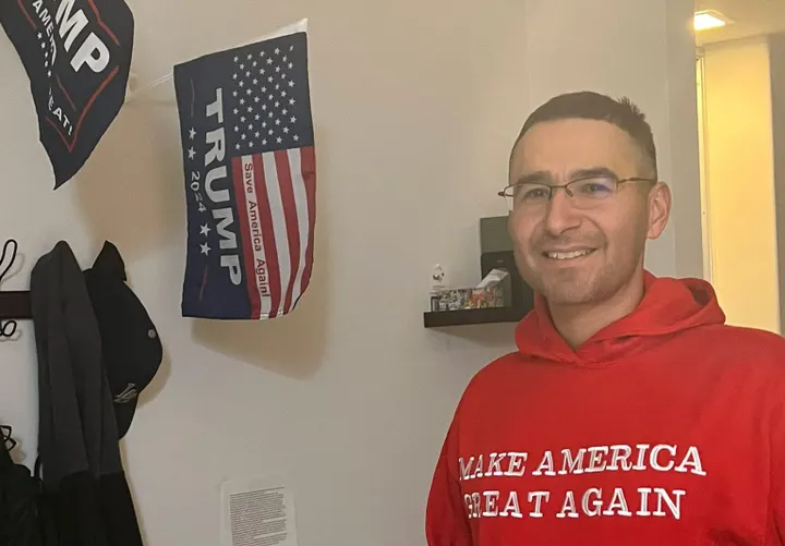 Man wearing a red “Make America Great Again” hoodie stands indoors, smiling, with two pro-Trump flags hanging on the wall behind him.
