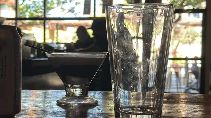 A small martini-style glass with a dark layered drink sits on a wooden table beside an empty pint glass, with sunlight streaming through large windows in the background.