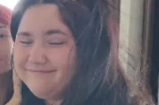 A 12-year-old girl with long dark brown hair smiles softly with her eyes closed. She is wearing a black top and appears indoors.