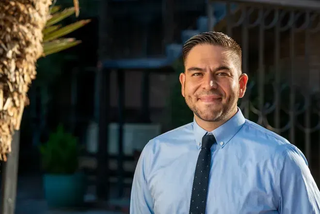 Rep. Gabe Vasquez stands outdoors in a light blue shirt and dark tie, smiling at the camera.