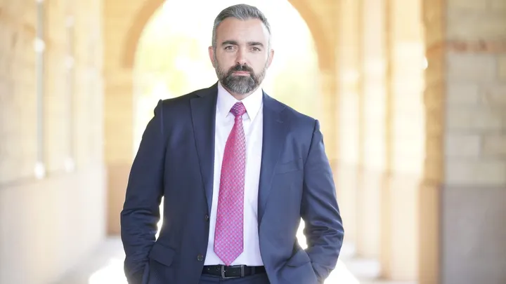 New Mexico Attorney General Raúl Torrez stands in a suit and tie under an arched walkway, looking directly at the camera.