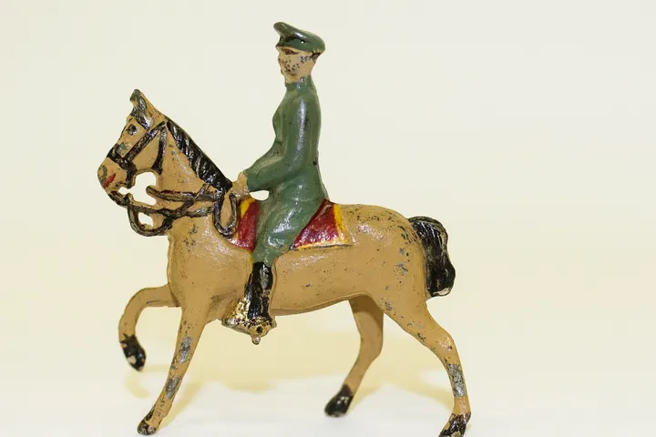 ChatGPT said:  A vintage cast-metal toy soldier figurine in a green uniform and cap rides a tan horse with a red saddle, shown against a plain light background.