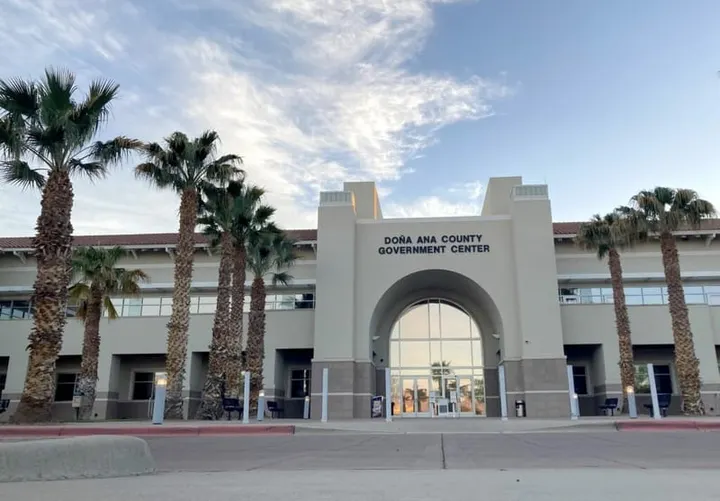 The Doña Ana County Government Center in Las Cruces, shown from the front entrance with palm trees lining the walkway under a partly cloudy sky.