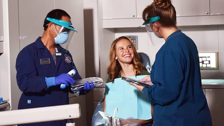 A patient smiles while talking with two masked dental professionals preparing instruments at Doña Ana Community College’s dental clinic.