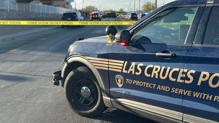 Las Cruces Police Department vehicle blocks a street behind yellow crime scene tape as officers investigate in the distance on a sunny afternoon.