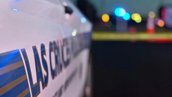 Close-up of a Las Cruces Police Department vehicle with blurred flashing lights and yellow crime scene tape in the background.