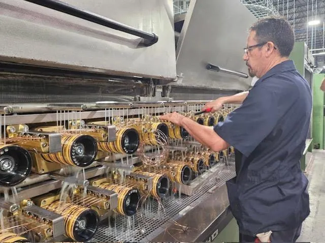 A worker operates machinery at a manufacturing facility, adjusting multiple spools of copper wire as the equipment runs.