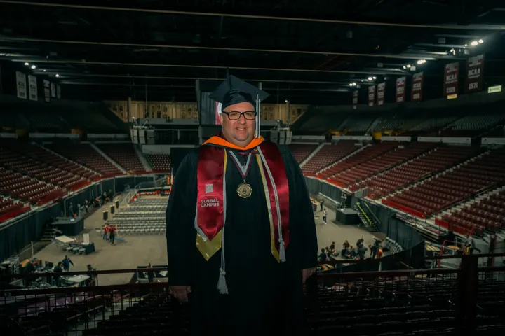 NMSU Global Campus graduate stands in cap and gown inside the Pan American Center after commencement.