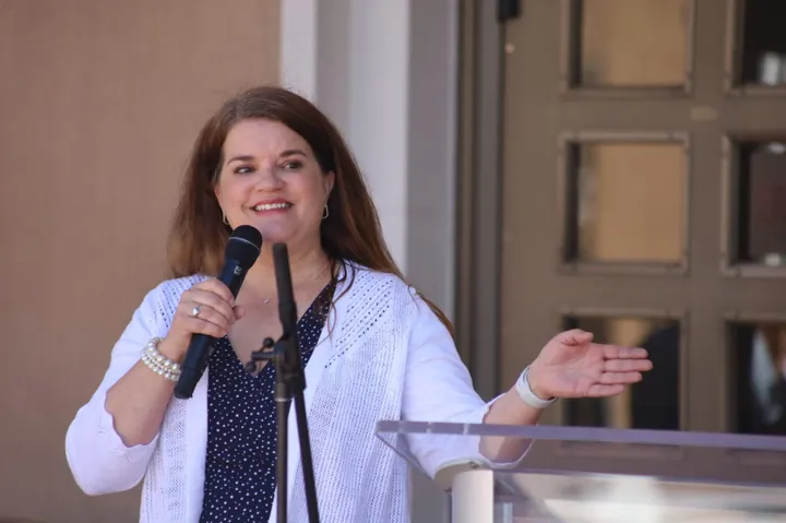 New Mexico Secretary of State Maggie Toulouse Oliver speaks into a microphone and gestures with one hand while standing at a podium during a public event outside a government building.