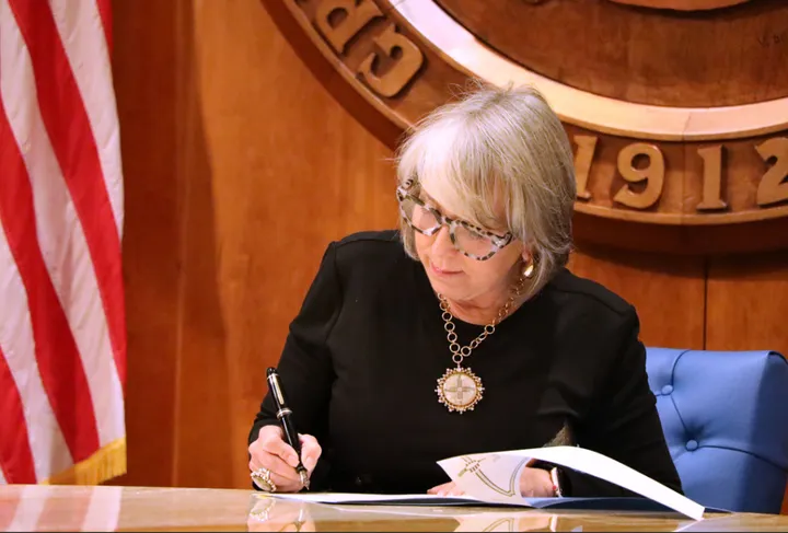 New Mexico Gov. Michelle Lujan Grisham signs legislation at her desk inside the Roundhouse, seated beneath the state seal with an American flag in the background.