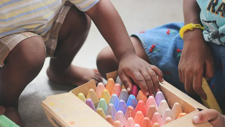 Two young children sit on the floor playing with a box of colorful sidewalk chalk, their hands reaching for pieces during an early learning or preschool activity.