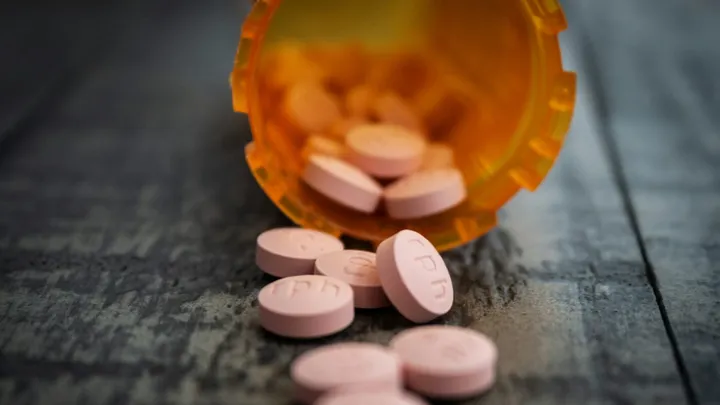 An open orange prescription pill bottle tipped over on a wooden surface with pink tablets scattered in front of it.