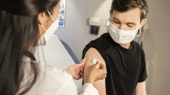 A masked health care worker applies a bandage to a patient’s upper arm after administering a vaccination.