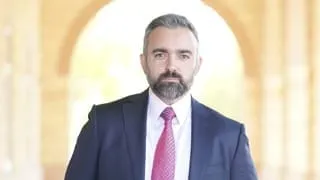 Attorney General Raúl Torrez, in a navy suit, white shirt and pink tie stands facing the camera inside a softly lit arched corridor with a bright background.