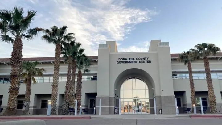 Exterior of the Doña Ana County Government Center in Las Cruces showing its arched glass entrance, beige facade and rows of palm trees under a partly cloudy sky.