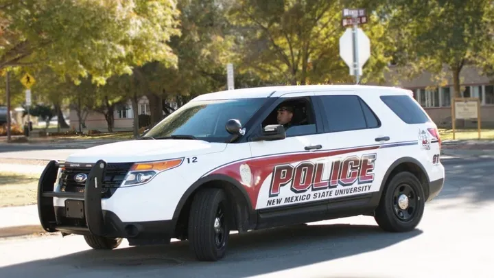 A New Mexico State University police SUV drives through campus as an officer looks out from the driver’s seat, with trees and campus buildings in the background.