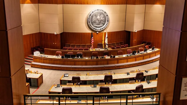 The New Mexico State Senate chamber in Santa Fe, showing rows of empty desks, maroon chairs, and the state seal mounted on the wall behind the podium.