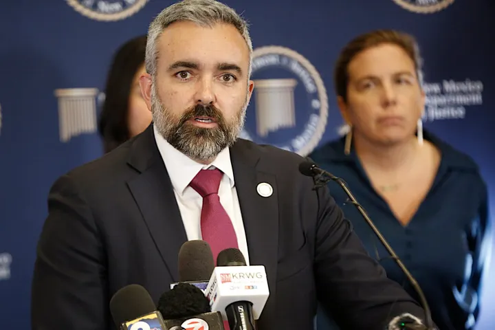 New Mexico Attorney General Raúl Torrez speaks at a press briefing behind microphones, wearing a dark suit and red tie, with a blurred Department of Justice backdrop and staff behind him.