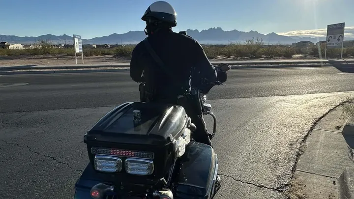 A Las Cruces police officer on a motorcycle watches traffic on Red Hawk Golf Road with the Organ Mountains visible in the distance.