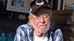 A 68-year-old man wearing a dark baseball cap, glasses, and a blue plaid shirt sits indoors with his arms crossed. He has gray facial hair and a serious expression.